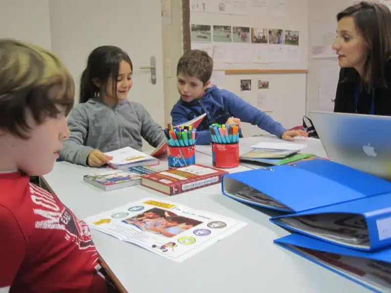 **Alt Text:** A teacher and students sitting around a table, engaged in a group activity.