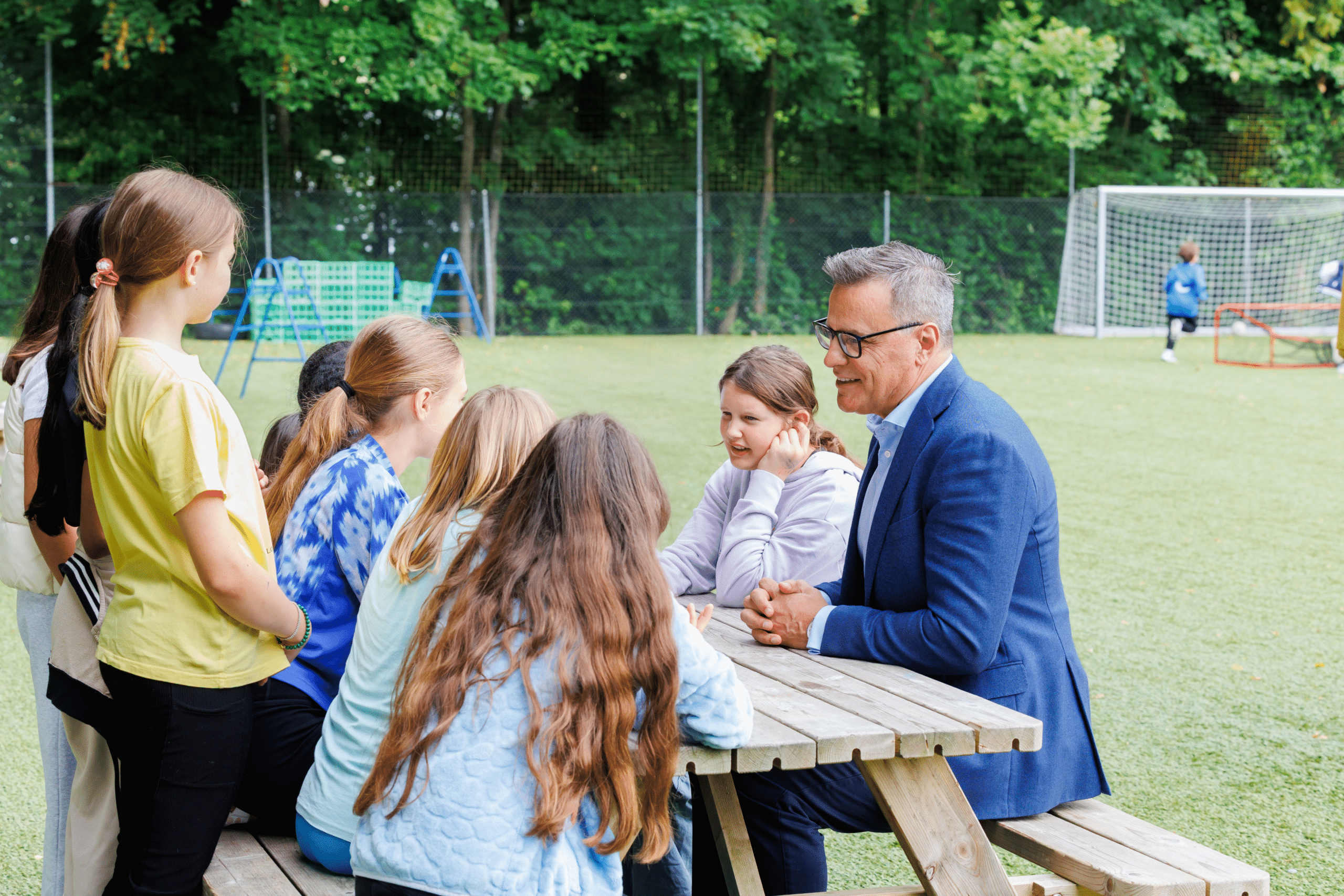 A middle aged man sitting at a wooden picnic table with a group of young Middle School students. In the background is a green football pitch.
