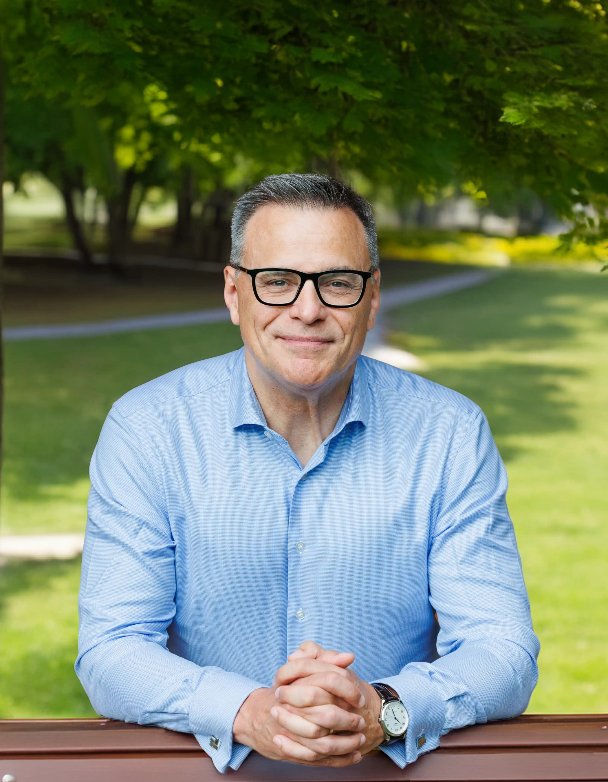 ISZL School Director, wearing glasses and a light blue shirt, stands outdoors with his hands resting on a wooden railing.