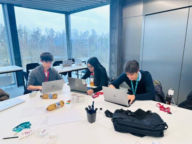 Three students sit around a table in a bright meeting room with large windows, working on laptops. Papers, notebooks, drinks, and bags are spread across the table as they collaborate on a project.
