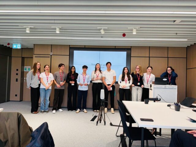 A group of students wearing red lanyards stand at the front of a modern conference room, presenting in front of a large screen. A tripod with a recording device is set up in front of them, while tables and chairs are arranged in the foreground.