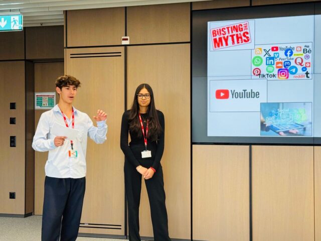 Two students wearing red lanyards present at the front of a conference room. One student speaks while holding notes, and a slide behind them reads “Busting the Myths” alongside social media logos including YouTube, TikTok, and Instagram.