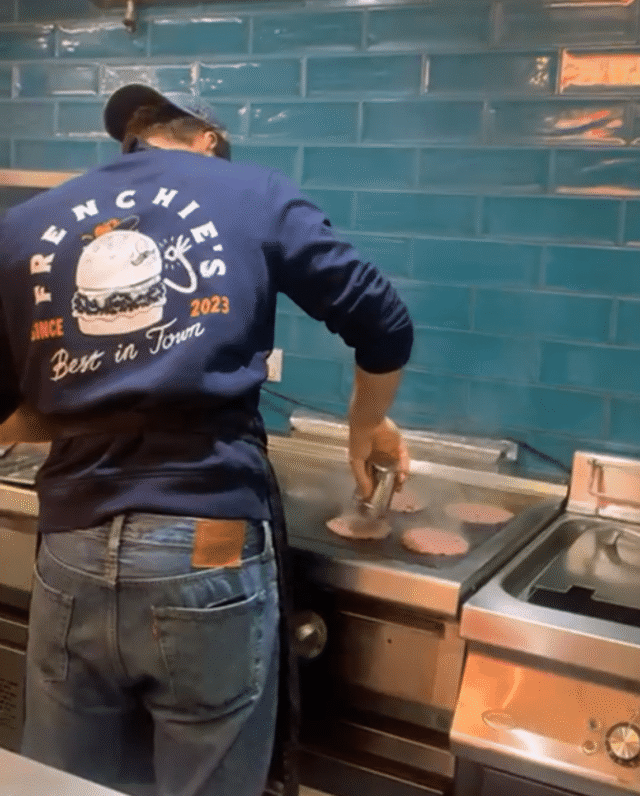 A chef stands at a flat-top grill in a restaurant kitchen, pressing burger patties while wearing a navy sweatshirt with “Frenchies – Best in Town” printed on the back.