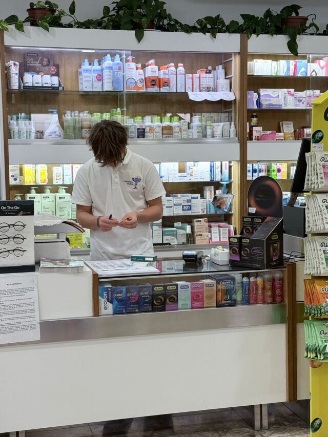 A student wearing safety glasses works at a laboratory bench, carefully mixing a solution in a small beaker beside trays of sample containers and lab equipment.