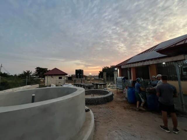Sunset over a community site with large circular water tanks and solar panels, as several people work together beside a building.