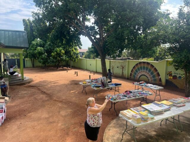 Open courtyard with folding tables covered in books and school supplies, as a woman takes a photo and others stand nearby under trees.