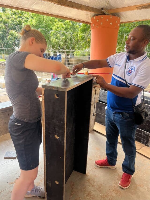 A young woman and a man measure and assemble a tall wooden cabinet or box under a covered outdoor space.