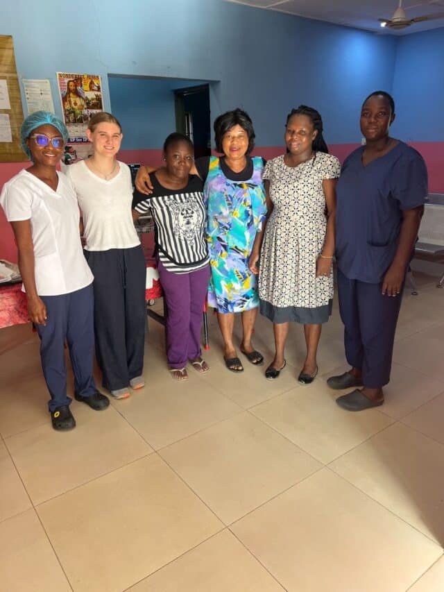 Six women standing together indoors, smiling for a group photo in what appears to be a clinic or community space.
