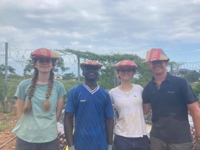 Four people standing side by side at a construction site, smiling and balancing woven hats on their heads.