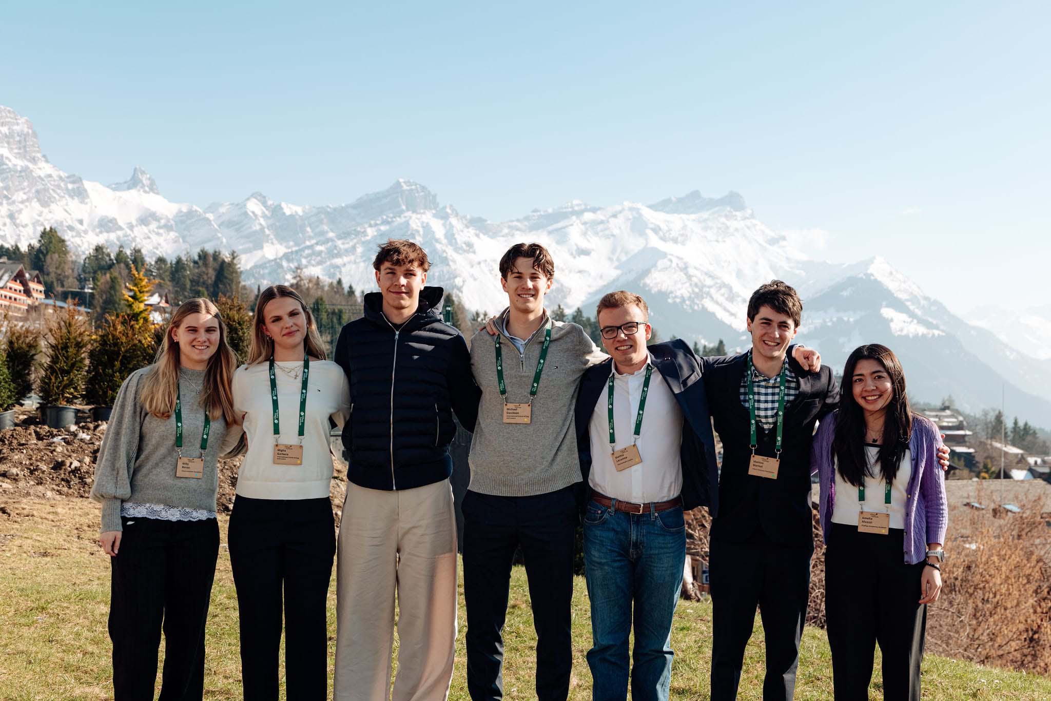 Seven students stand arm in arm outdoors wearing conference badges, with snow-capped mountains and alpine scenery in the background.