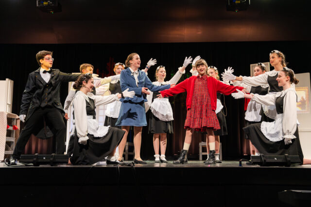 A group of students perform a musical number on stage, with a central performer in a red dress and cardigan surrounded by others in black-and-white costumes, all gesturing outward in a choreographed scene.
