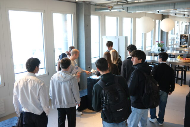 A group of students gather around a presenter in a bright, modern space, listening as a demonstration is shown on a small display table.