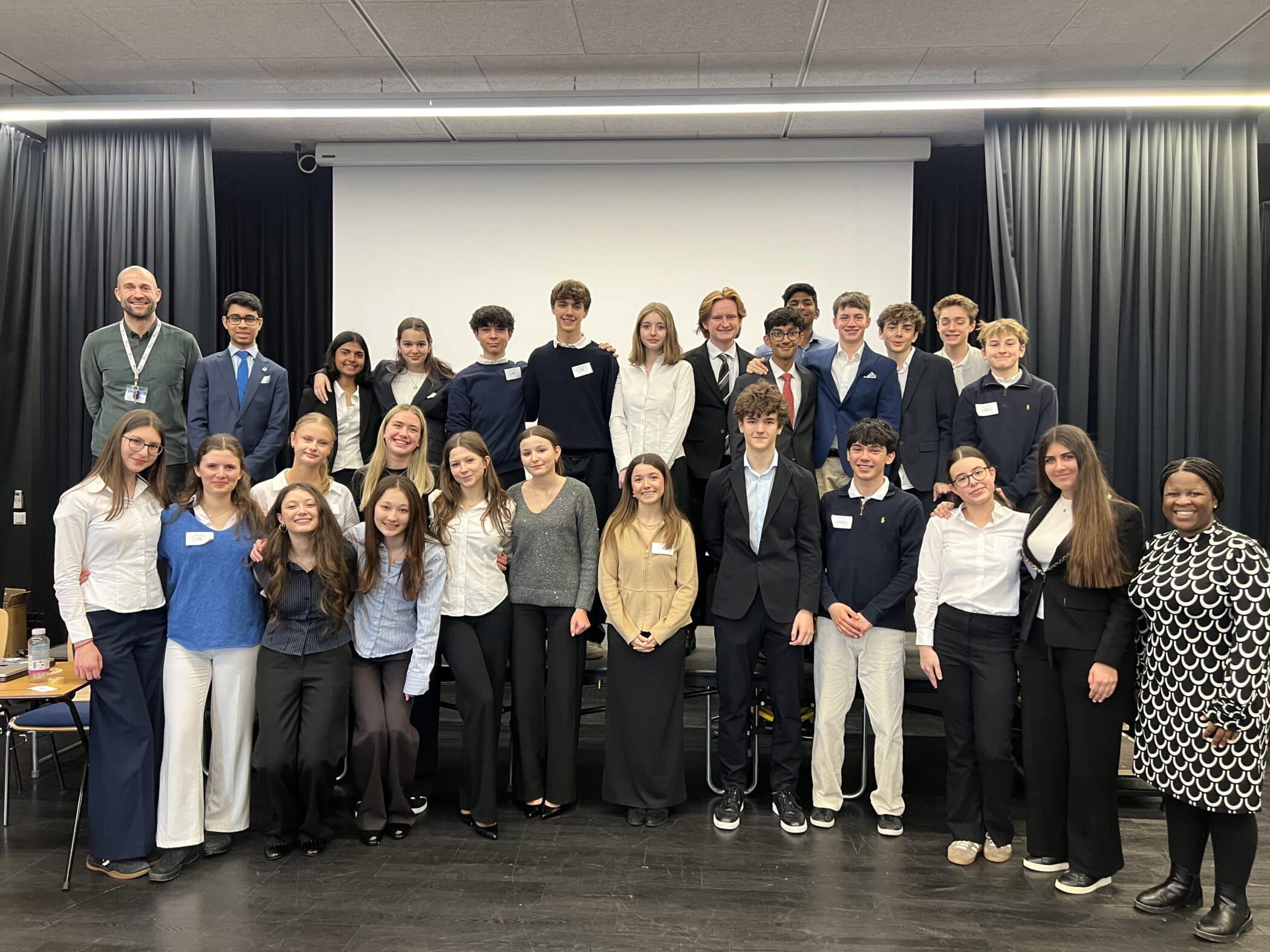 A group of students and teachers stand together for a group photo at the end of the ISZL Model United Nations conference, posing in front of a presentation screen in a conference room.