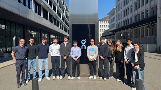 A group of students and staff stand outdoors in front of a modern building, gathered around an On running brand sign, posing together in a sunny courtyard.