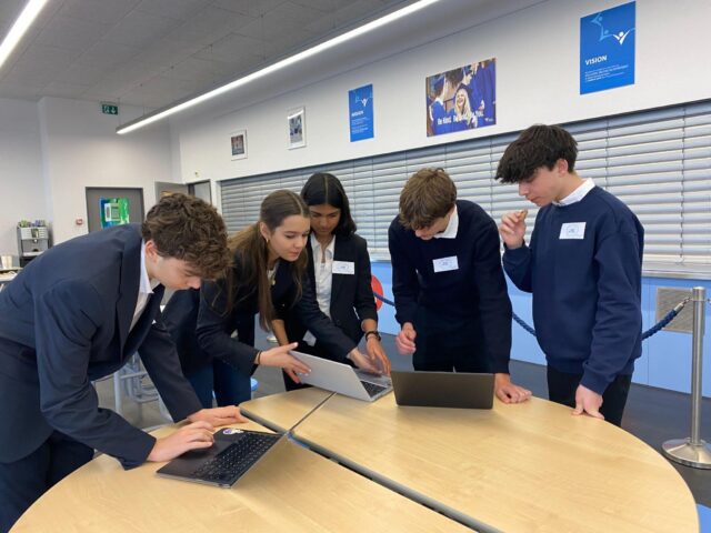 Five students stand around a table working on laptops together, leaning in to collaborate on a task in a bright school space.