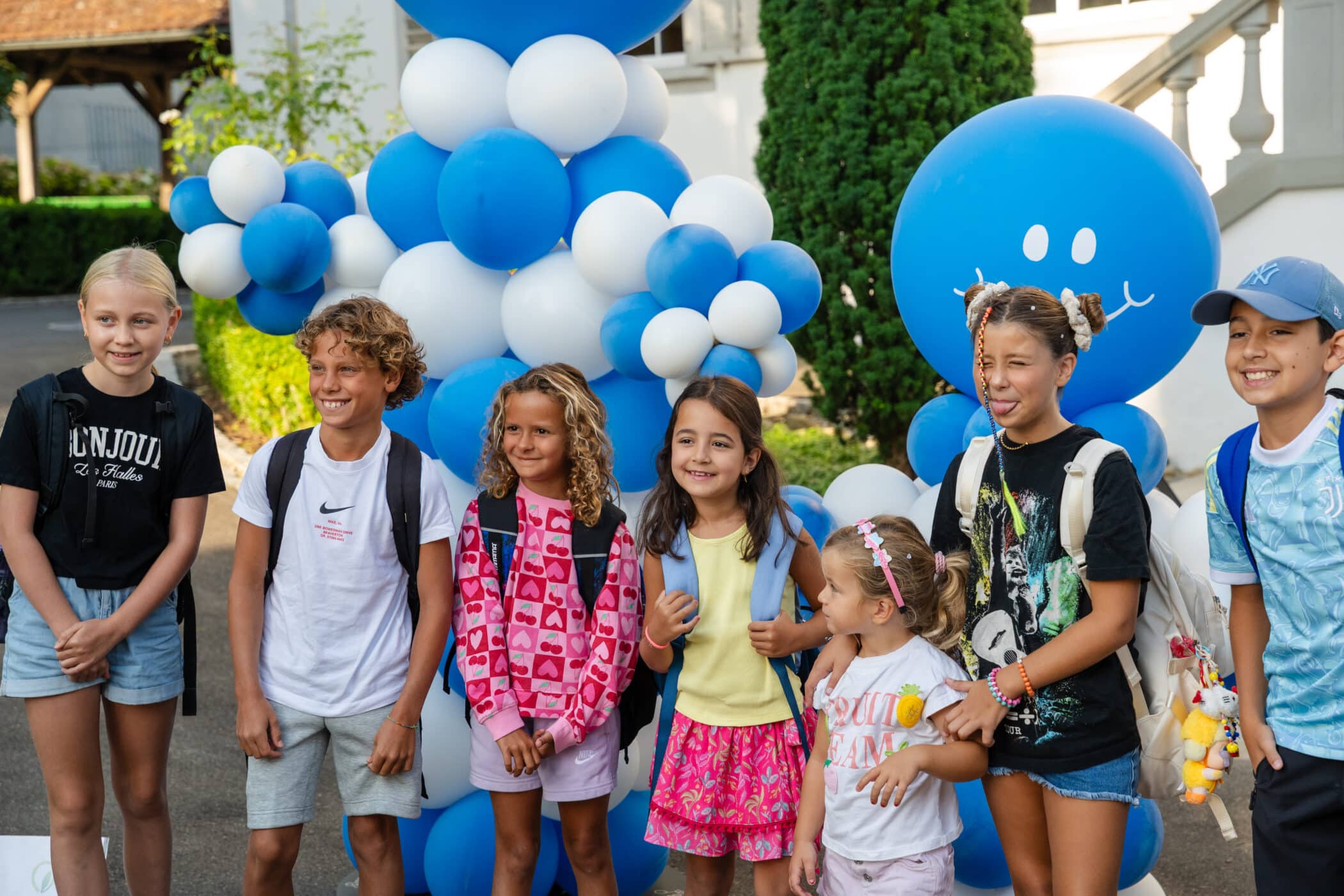 A group of children wearing backpacks stand smiling in front of large blue and white balloon decorations shaped with friendly faces.