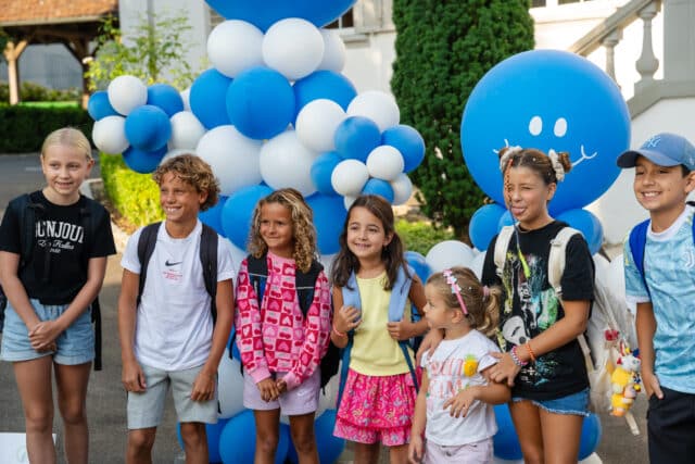A group of children wearing backpacks stand smiling in front of large blue and white balloon decorations shaped with friendly faces.