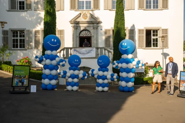 The entrance of a school building is decorated with blue and white balloon displays, as two adults walk past and signage welcomes students back.