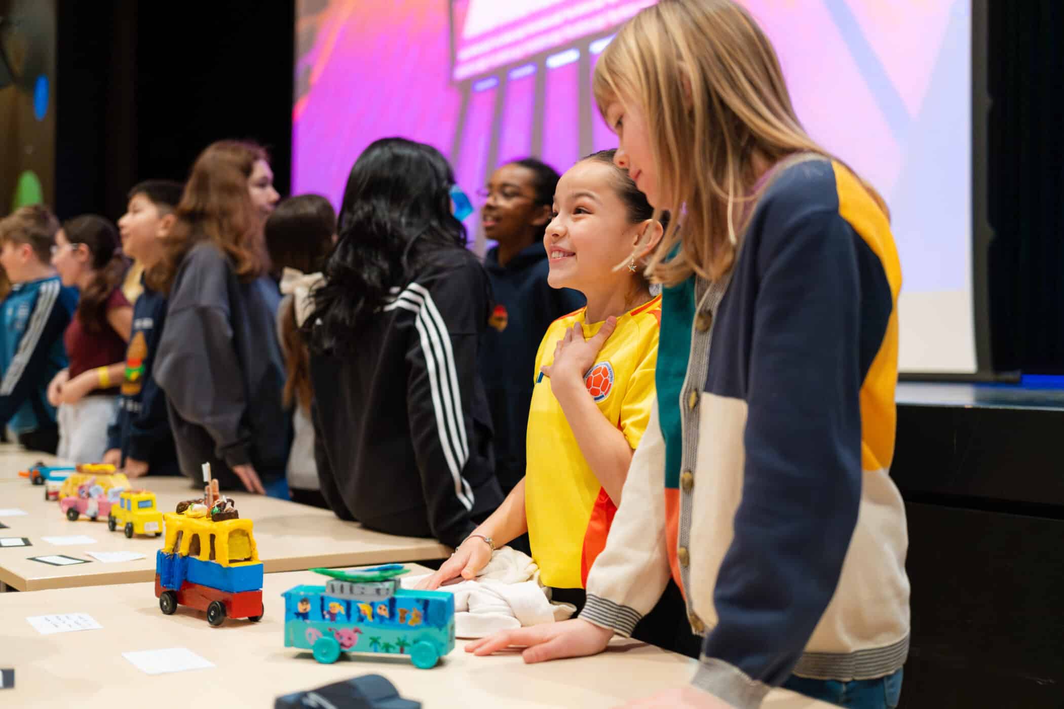 Students stand beside a table displaying colourful handmade model cars, with one student speaking animatedly while others listen during a classroom showcase.