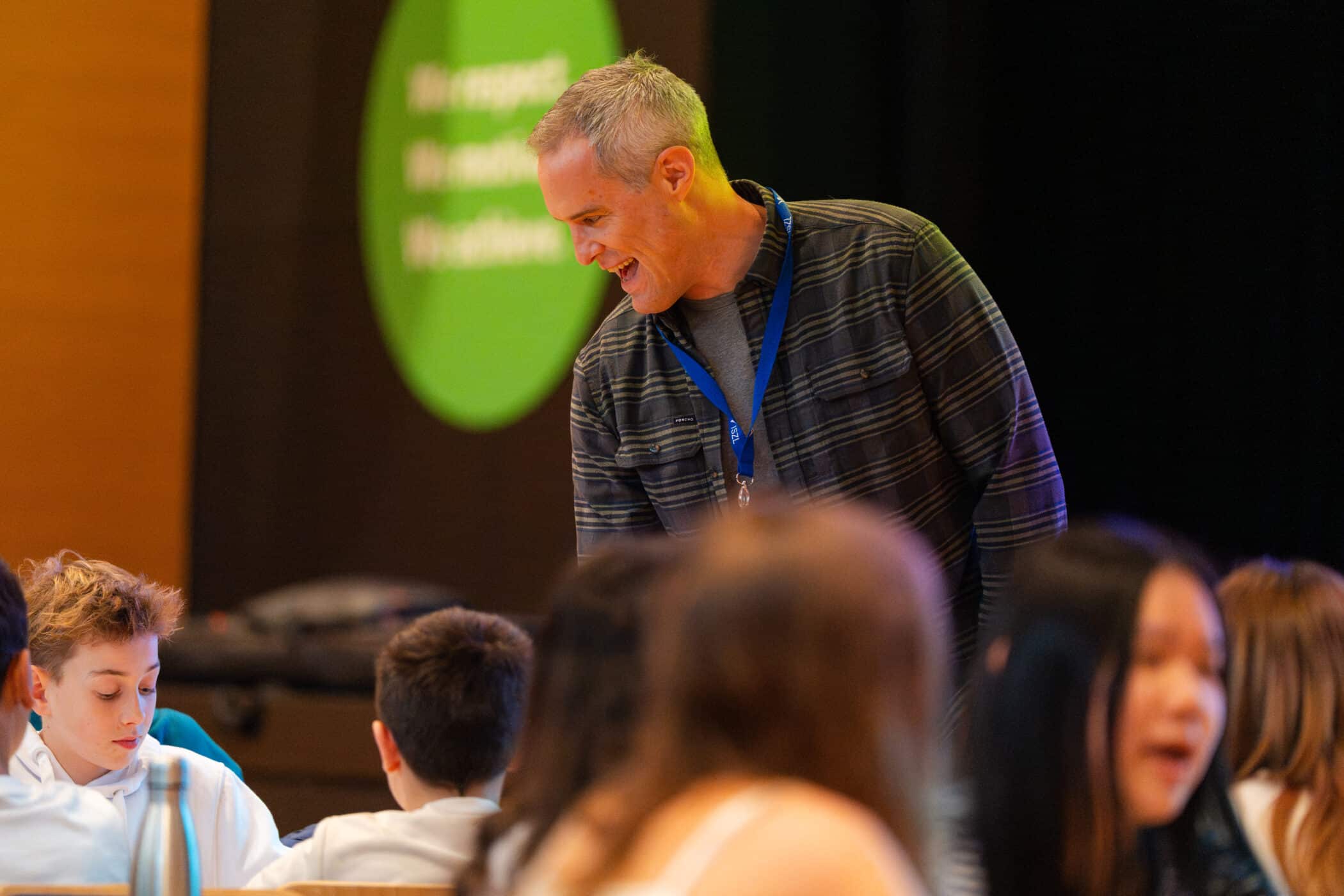 An author smiles and speaks with students gathered around tables, engaging with them during a school visit or workshop.