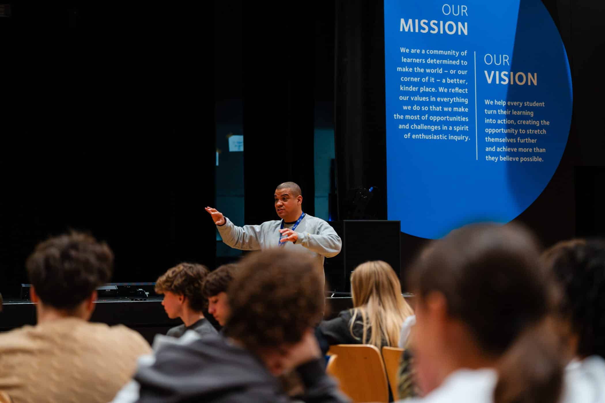 A speaker gestures while presenting to an audience of students in a theatre, with a large screen behind displaying the school’s mission and vision statements.