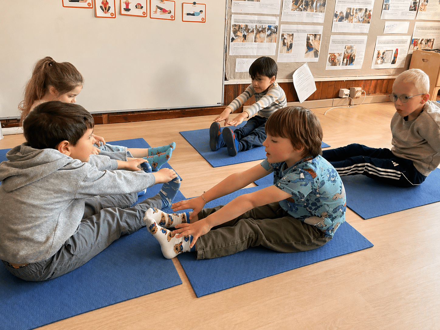 Young students sit on yoga mats in a classroom, stretching forward to touch their toes during a group physical activity session.