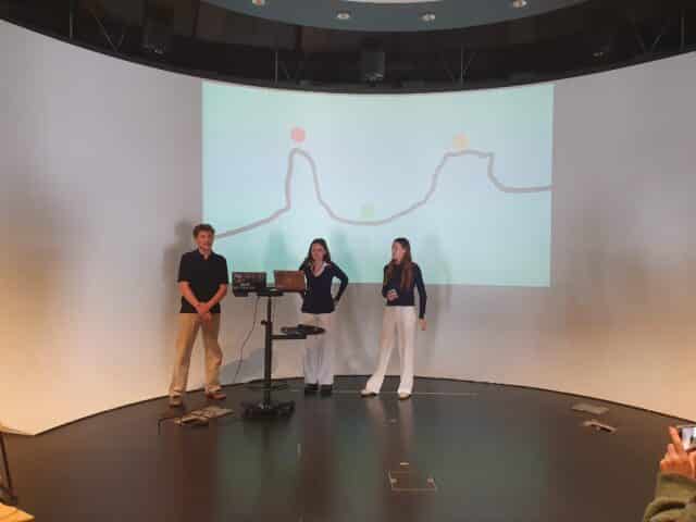 Three students stand beside a laptop presenting an interactive visual project on a large curved screen in a modern presentation space.