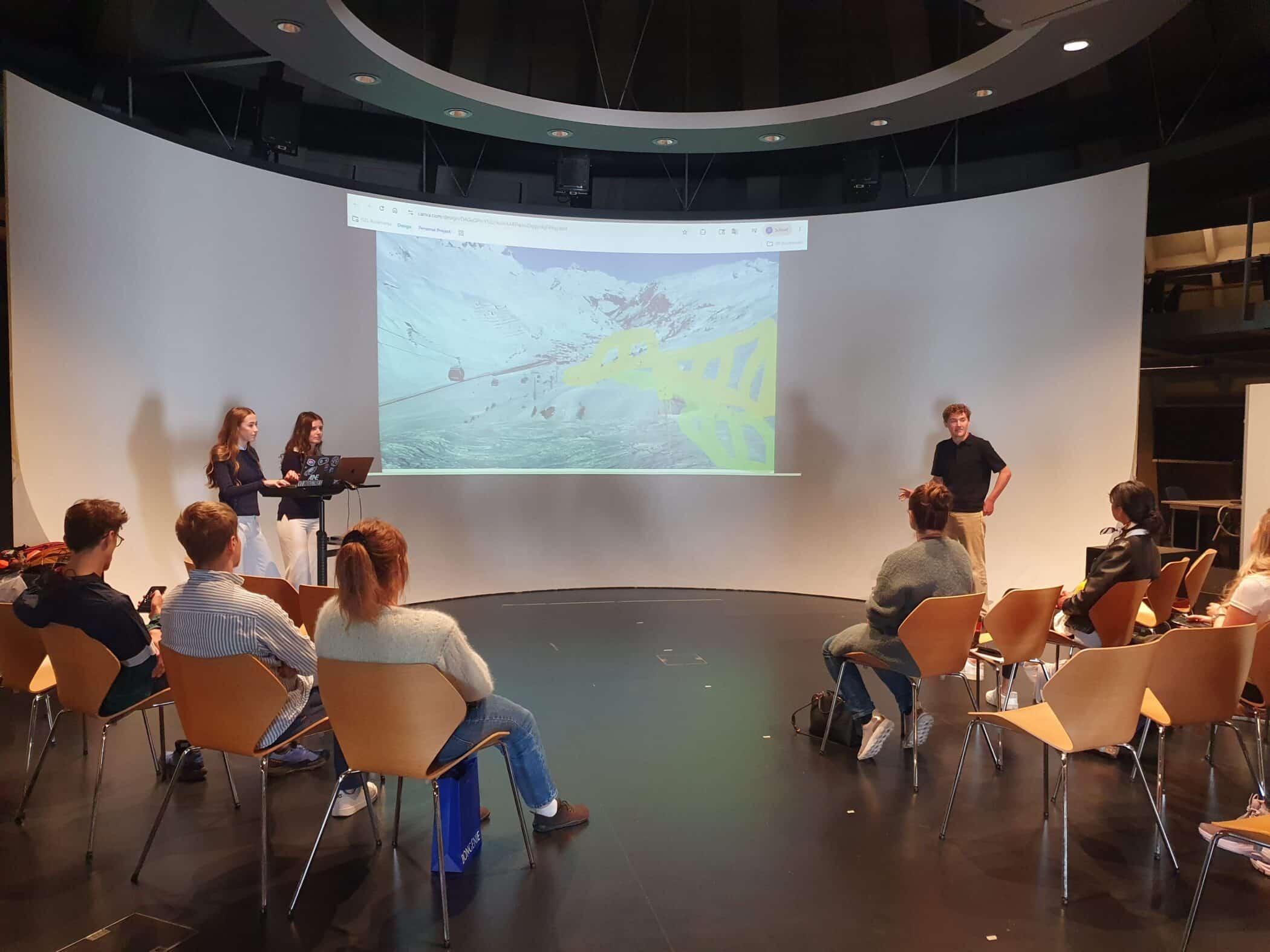 Students present a project to a seated audience, with a large projected image displayed across the curved wall behind them.