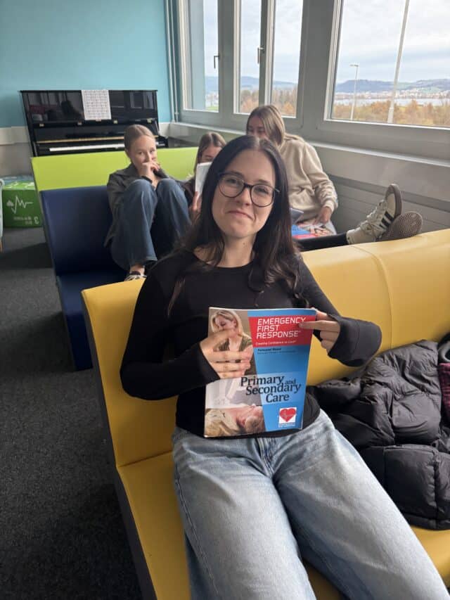 A student sits in a learning space holding a first aid training manual while classmates study in the background.