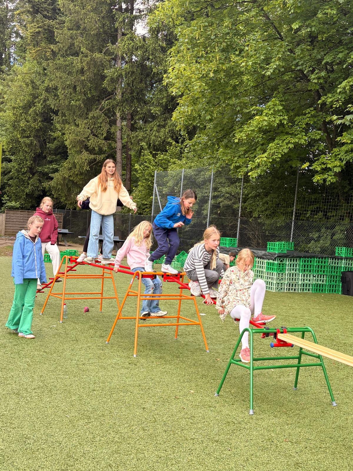 Children balance and climb on outdoor agility equipment set up on a sports field, working together during a playground activity surrounded by trees.