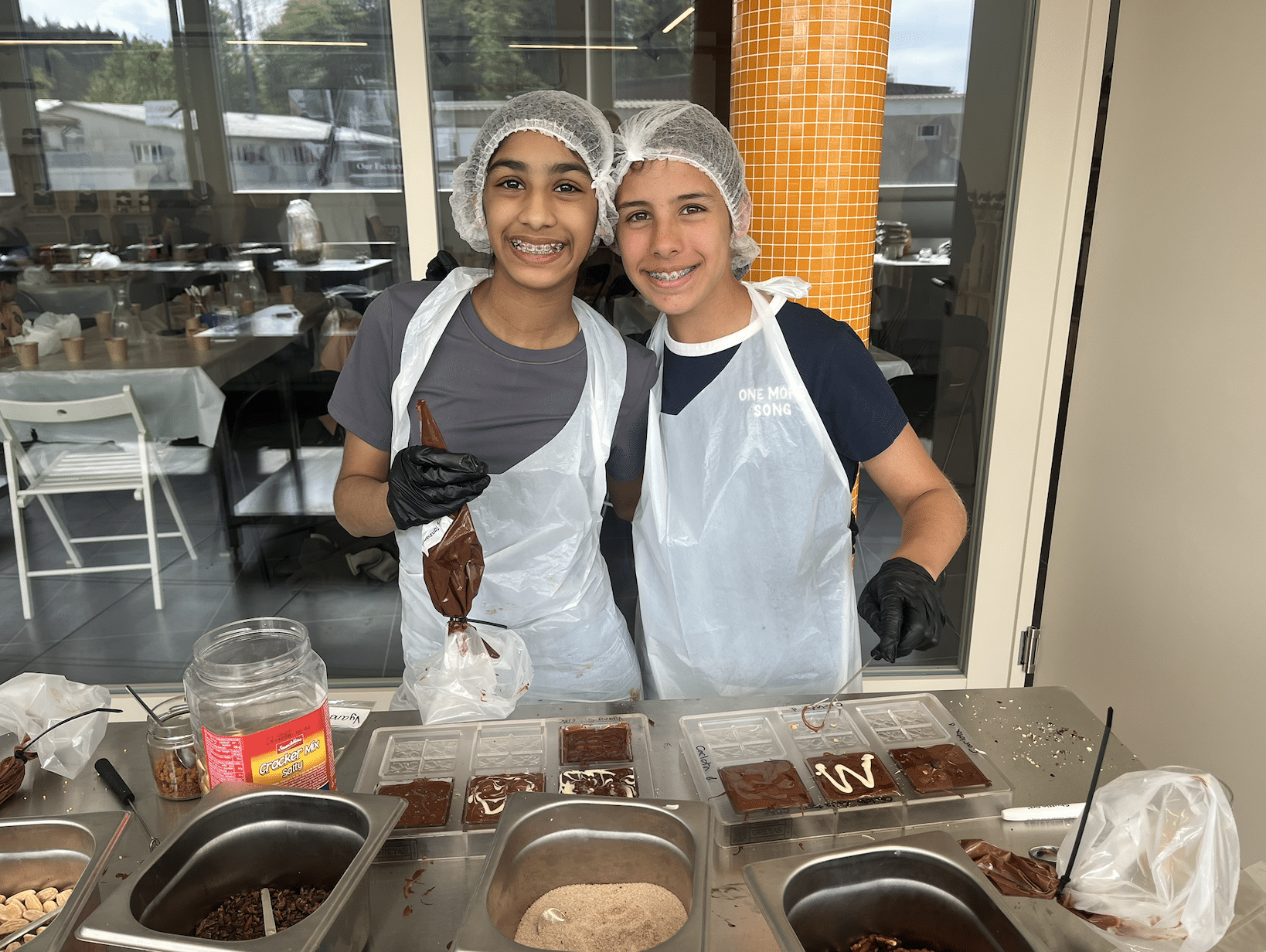 Two students wearing hairnets, aprons, and gloves smile at the camera while decorating chocolate bars at a workshop table.