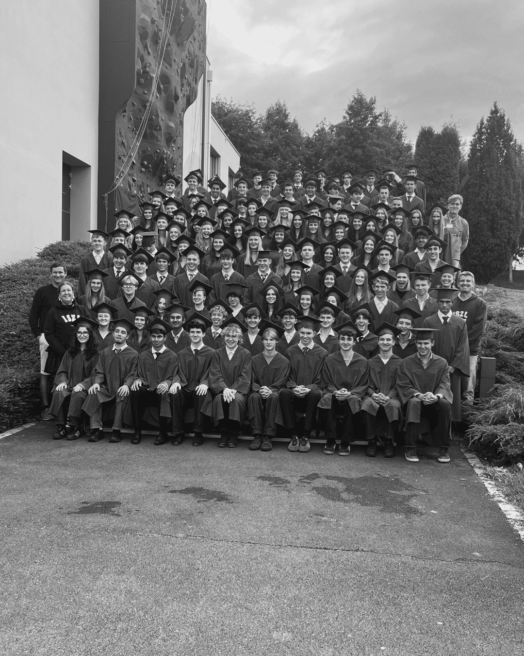 Class of 2026 graduation photo: rows of ISZL graduates in caps and gowns seated and standing on outdoor steps, with faculty and staff standing alongside them, in front of the school building and climbing wall.